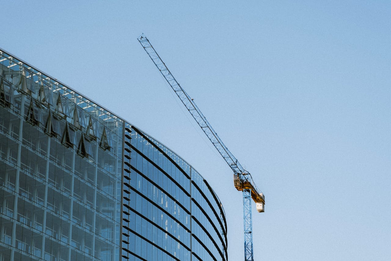 why-choose-us Contemporary glass building under construction with a visible crane against a clear blue sky.